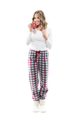 Wall Mural - Joyful cute young woman drinking coffee smiling at camera in cozy pajama and warm socks. Full length portrait on white background.