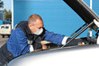 © SerPak - A white man wearing a medical mask on his face repairs a car outside in an open space. A mechanic or car mechanic in work clothes repairs or checks the operation of the car with the hood open.