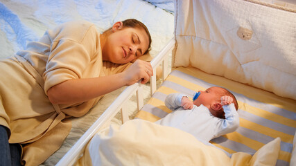 young tired mother fallen asleep while rocking her baby son in crib at late night. motherhood and sl