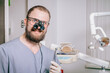 © indigo_nifght - A young dentist with a beard and a microscope on his glasses, holding a drill in his hands.