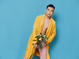 Asian man in a yellow coat on a blue background with a bouquet of flowers bent to the side