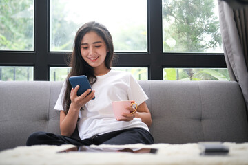 Happy young woman sitting on sofa and using mobile phone.