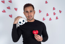 Young Man Holding A Red Love Heart On His Chest And A Baby Seal Plush On His Shoulder, With Red Paper Hearts Flying Behind Him. Concept Of Love, Valentine 