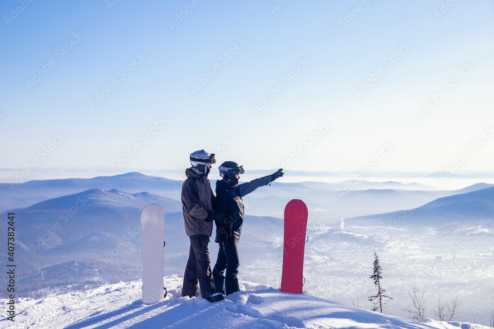 Loving couple of snowboarders hold hands and look forward, man indicates direction to woman with snowboard route with hand - obrazy, fototapety, plakaty 