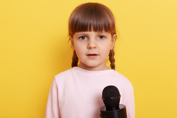 Cute shy child wearing pale pink shirt speaking in microphone, looks at camera with a bit confused look, small kid with pigtails being interviewed against yellow wall.