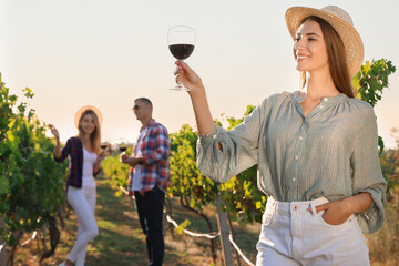 Wall Mural - Beautiful young woman with glass of wine and her friends in vineyard on sunny day