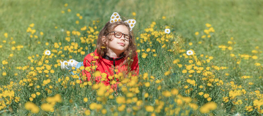 Portrait of funny brunette little girl with red glasses, curly windy hair smiling, closing eyes with satisfied expression, posing for family photo into green meadow with  dandelions. Banner.