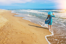 Seascape On A Sunny Summer Day. The Girl On The Beach Looks At The Sea. Young Happy Girl Carefree Walk Along The Sea In Blue Fluttering Dress And Sun Hat