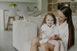 © VK Studio - Beautiful brunette affectionate mother holds small curly haired cute dauhter in dressing gown pose in cozy room against modern apartment interior. Mom and little girl at home after taking shower