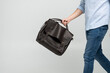 Leinwandbild Motiv Brown men's shoulder leather bag for a documents and laptop holds by man in a blue shirt and jeans with a white background. Satchel, mens leather handmade briefcase.