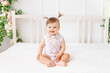 © Any Grant - cheerful baby girl six months old sitting in a bright beautiful room on a white bed in a lace bodysuit and smiling