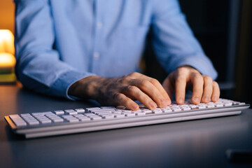Close-up view of hands of unrecognizable man working typing on wireless keyboard computer. Male freelancer or student is working sitting on desk at home office at late night in dark room.