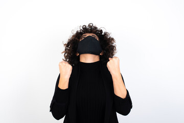 Wall Mural - young beautiful caucasian woman wearing medical mask standing against white wall looks with excitement up, keeps hands raised, notices something unexpected.