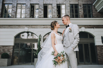 Newlyweds in love hug on the background of a brick multi-storey building with windows on the street in the city. Wedding portrait of the groom in a gray tailcoat and the bride in a white dress.