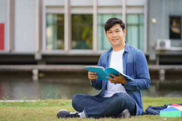 Portrait of Asian man university student aitting on grass in campus looking at camera and smile and reading a book in park..