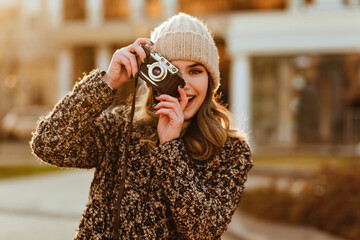 Poster - Blissful female model in elegant coat posing with camera. Smiling photographer taking pictures of autumn city.