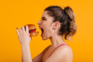 Studio shot of hungry woman with sandwich. Caucasian girl eats hamburger with pleasure.