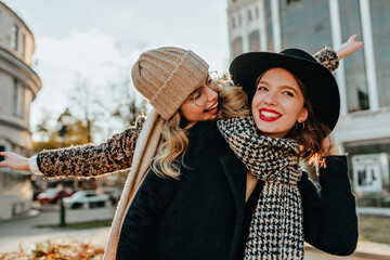 Wall Mural - Wonderful woman in checkered scarf having fun with friend. Outdoor portrait of two gorgeous girls enjoying autumn.