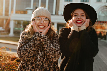 Excited woman with wavy hair posing in autumn day with friend. Outdoor photo of two pretty sisters standing on city background.