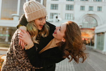 Wall Mural - Debonair girls enjoying autumn weekend. Pretty young woman in knitted hat embracing her friend.