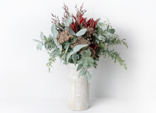 Beautiful Flower Arrangement Of Mostly Australian Native Flowers, Including Silvan Reds, Queen Anne's Lace, Wattle Foliage And Eucalyptus Leaves, In A Ceramic White Vase, With A White Background.