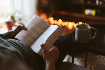 A book in man's hand, a man sitting on a couch with a cup of tea, warm candid light in a cosy living room