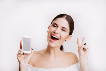 Sticker - Closeup portrait of brunette girl with phone in her hands. Young woman shows peace sign on white background