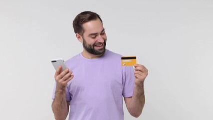 Wall Mural - Smiling pensive joyful bearded young man 20s in casual violet t-shirt isolated on white background studio. People lifestyle concept. Using mobile cell phone hold credit bank card doing winner gesture