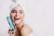 Satisfied female model without makeup smiling on white background. Girl after shower posing with bottle of cosmetic tonic
