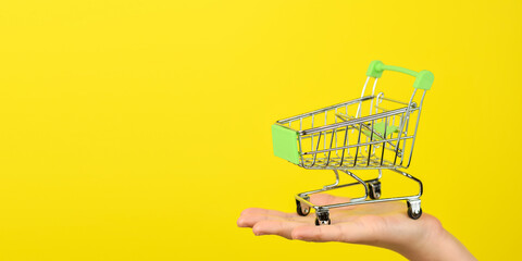 Empty shopping cart on female hand. Shopping theme. Small Trolley isolated on a bright yellow background. Consumer concept, Copy space.