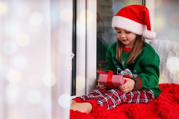 Poster - Cute little girl in Santa hat holding Christmas present on window sill at home