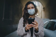 © Roman - Portrait of young dark-haired woman wearing mask and light-colored coat. She walks city streets and uses smartphone to chat with her friends. Good vibes. Modern technology advantages concept