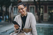© Roman - Young beautiful dark-haired Caucasian woman wearing glasses, sitting at the fountain, holding smartphone and smiling sweetly. Good vibes and feelings. Online communication using mobile digital device