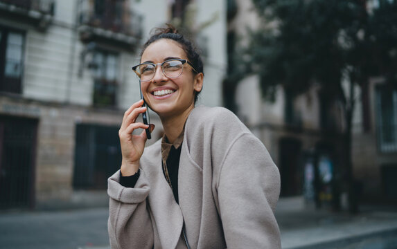 Portrait of young good-looking Caucasian woman wearing glasses, talking on her smartphone outdoors and smiling sweetly. Mobile digital devices make communication and interaction between people easier