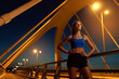 © Roman - Young female runner confidently standing with hands on hips on illuminated bridge at night and looking upwards. Athletic woman getting ready to exercise to maintain shape, weight and healthful mind