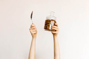 Woman holding a glass jar with granola and a spoon on white wall background. Healthy breakfast concept.