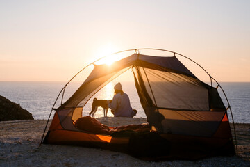 woman and her pet dog sit at beach next to tent, solitude camping in nature. sunset shot of young wo