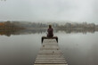 Calm morning relaxation by the foggy pond. Sitting woman in the countryside. Intended female mysterious atmosphere. Autumn afternoon in the nature. Silence. Wooden pier on a pond.