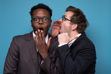 Caucasian man whispering to his african coworker ear telling him something secret. Studio shot on blue wall. Human emotions facial expressions