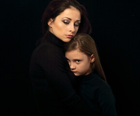 Wall Mural - Serious thinking mother hugging her sad daughter on black background in dark shadow. Closeup family emotional portrait