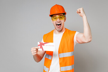 Poster - Young employee man in orange vest protective helmet hold gift voucher flyer do winner gesture clench fist isolated on grey background studio. Instruments for renovation apartment Repair home concept.