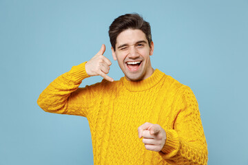 Young smiling friendly man 20s in casual knitted yellow fashionable sweater do phone gesture like says call me back point index finger camera on you blink isolated on blue background studio portrait