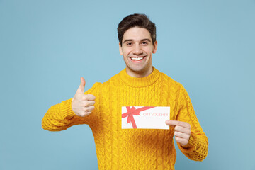 Young cheerful happy smiling student man 20s in casual knitted cozy yellow fashionable sweater hold in hand gift voucher show thumb up like gesture isolated on blue color background studio portrait