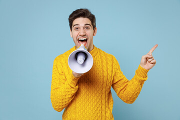 Young student man 20s in casual knitted yellow fashionable sweater scream aside shout in megaphone point index finger aside on copy space workspace area isolated on blue background studio portrait.