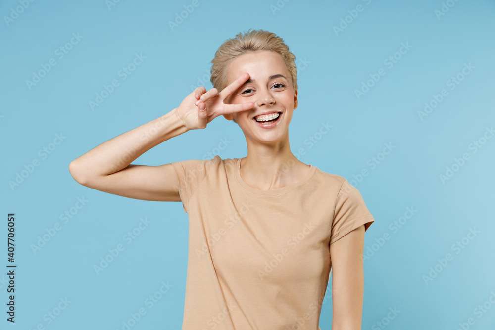 Young blonde smiling pretty caucasian woman dental braces short haircut nude make up in casual basic beige t-shirt showing v-sign victory gesture near eye isolated on blue background studio portrait