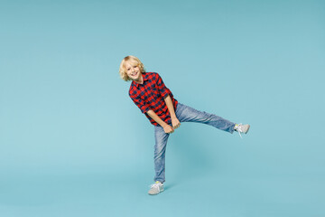 Full length of cheerful little curly kid boy 10s years old wearing basic red checkered shirt dancing rising leg isolated on blue color background children studio portrait. Childhood lifestyle concept.