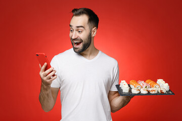 Wall Mural - Excited young man 20s in white t-shirt using mobile cell phone typing sms message hold makizushi sushi roll served on black plate traditional japanese food isolated on red background studio portrait.