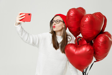 Pretty young woman in red hat glasses doing selfie shot on mobile phone blowing sending air kiss celebrating birthday holiday party hold heart air inflated helium balloon isolated on white background.