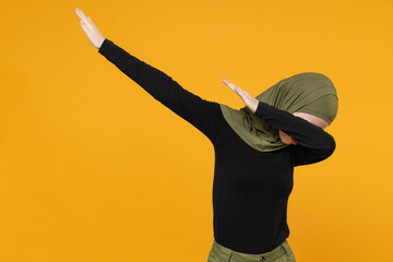 Wall Mural - Funny young arabian muslim woman in hijab black green clothes doing dab hip hop dance hands move gesture youth sign hiding cover face isolated on yellow background. People religious lifestyle concept.