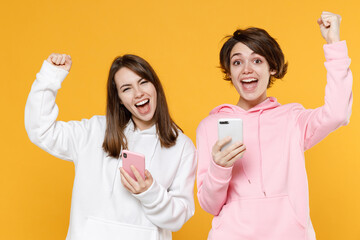 Joyful happy two young women friends 20s wearing casual white pink hoodies standing using mobile cell phone typing sms message doing winner gesture isolated on yellow color background studio portrait.